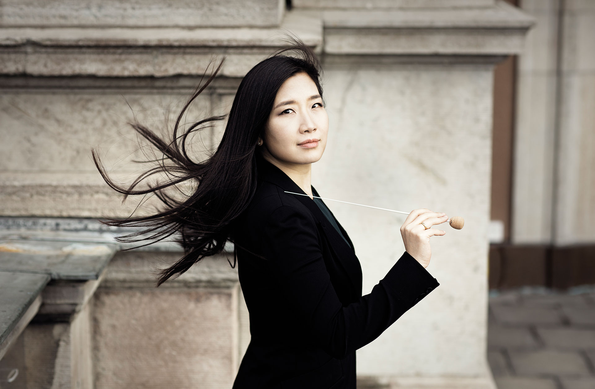 Kvinna med mörkt hår som flyger i vinden, med en taktpinne i handen.