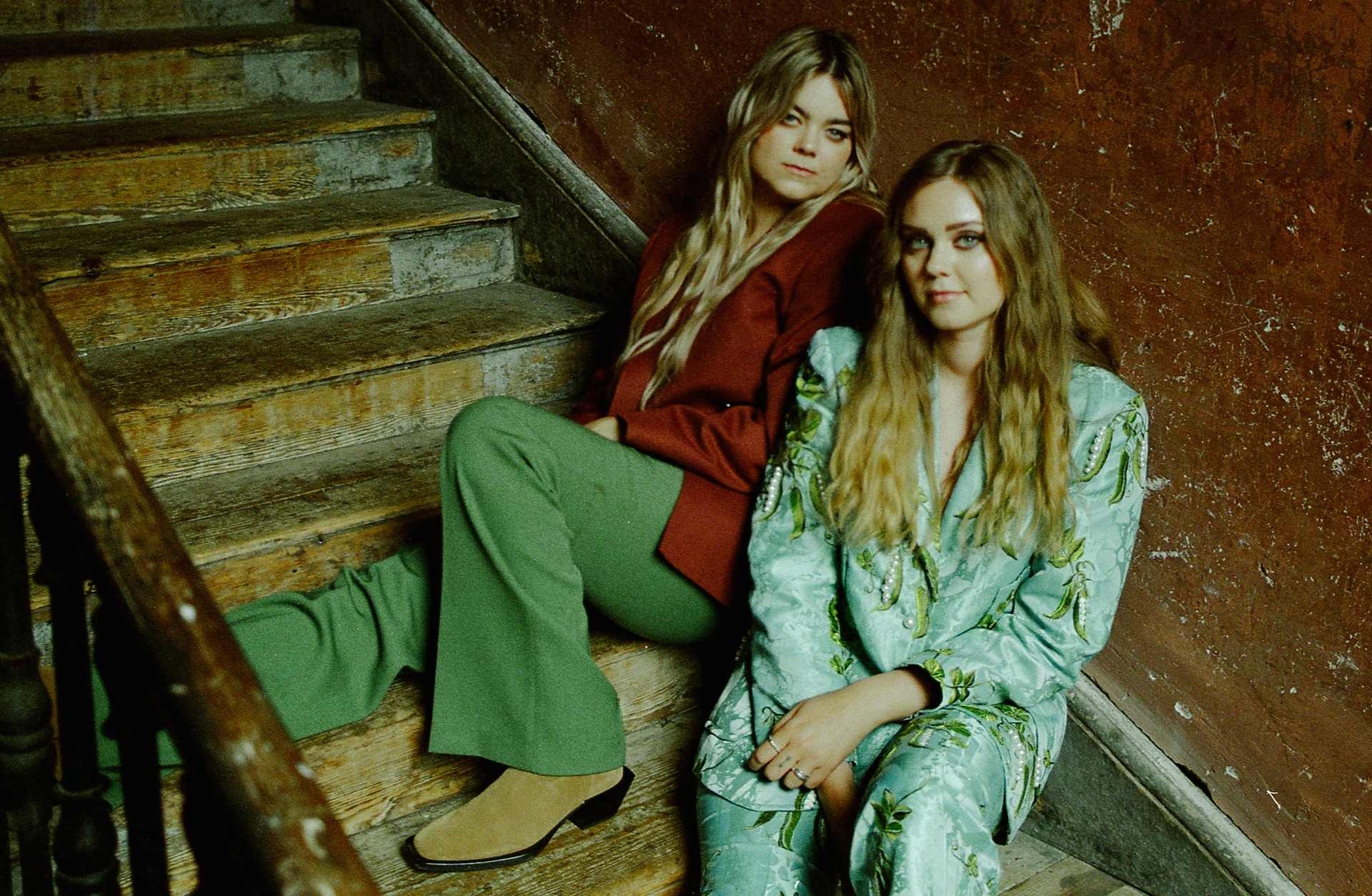 Två kvinnor sitter tillbakalutade i en trappa. De är blonda, med långt hår och de tittar in i kameran.