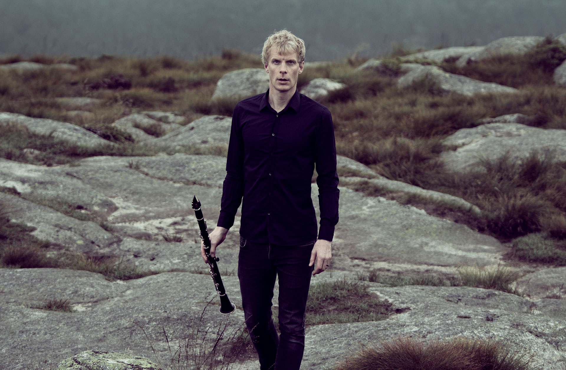 En svartklädd man med blont hår går på en berghäll med en klarinett i handen.