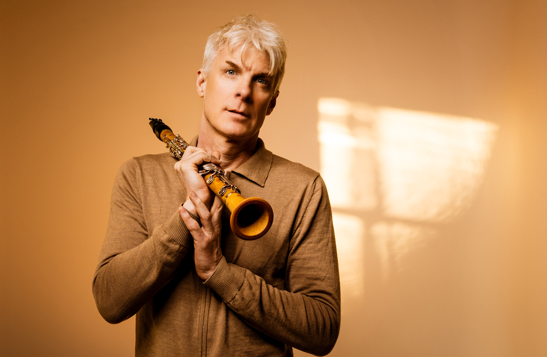 En vithårig man med en gyllenbrun klarinett står mot en gul vägg.