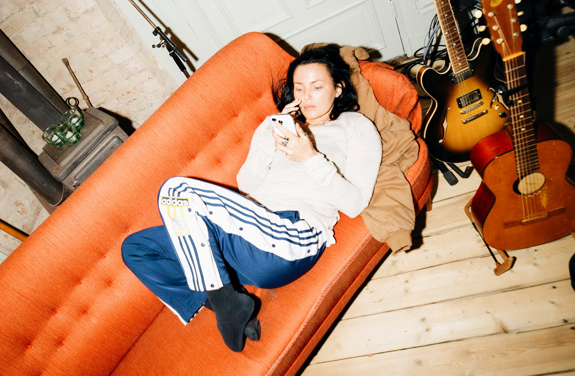 En kvinna med vit tröja och blåa träningsbyxor ligger och tittar i en mobil. Hon ligger på en orange soffa och har gitarrer bredvid sig.