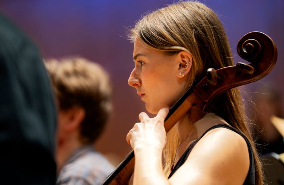 Närbild av en ung kvinna med långt hår som spelar cello med en orkester.