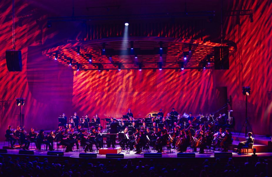 En orkester på en scen. De är belysta av ett rödlila ljus.