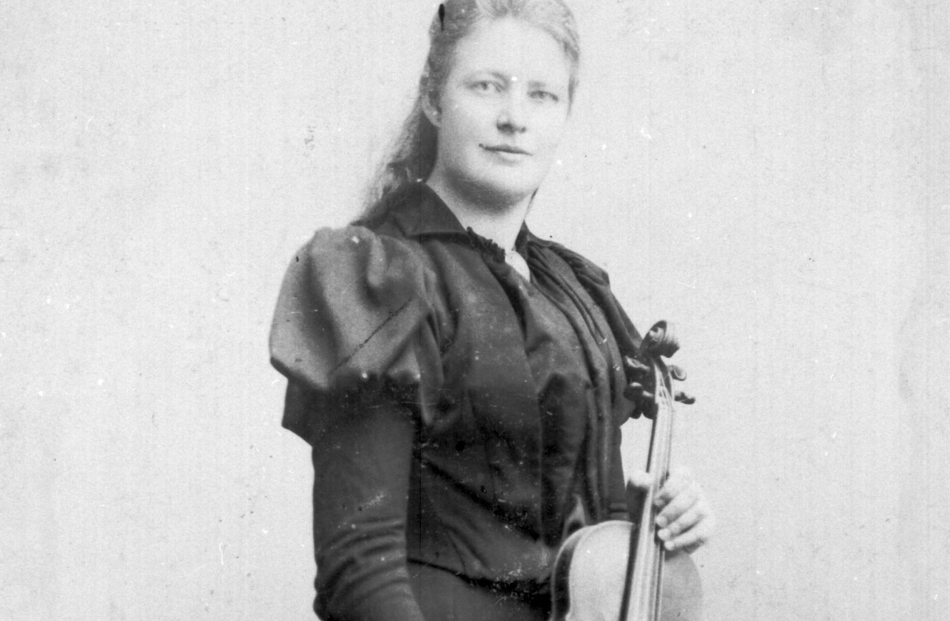 En svartvit historisk bild av en finklädd kvinna i klänning. Hon håller i en fiol.