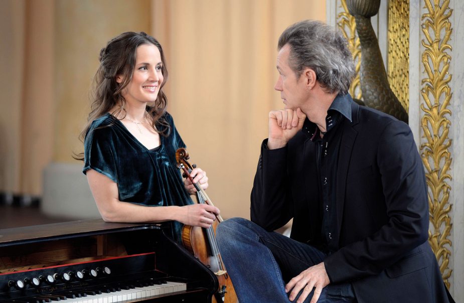 En kvinna håller en fiol i handen, lutar sig mot en orgel och tittar på en man med ryggen mot betraktaren.