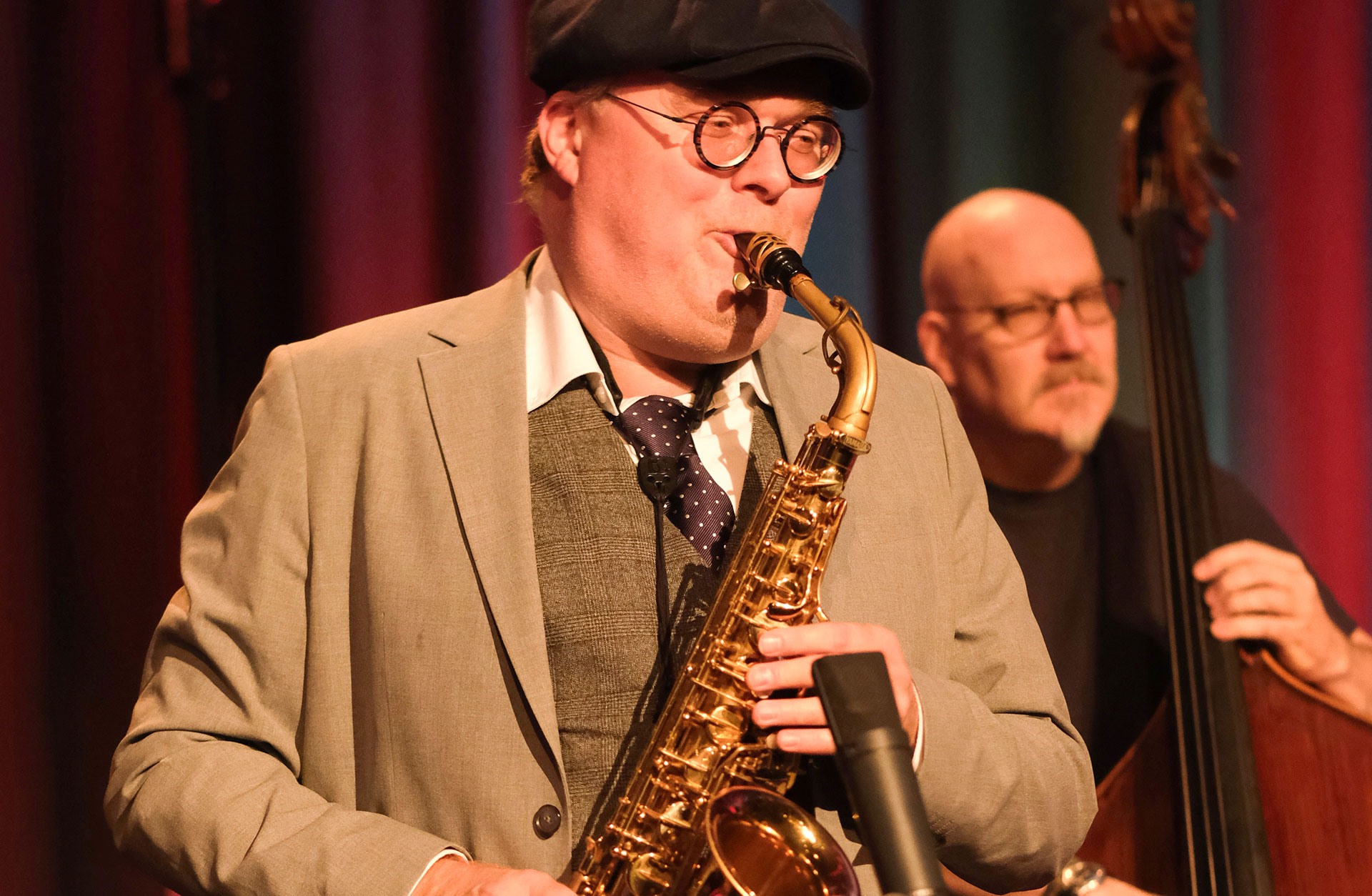 En äldre man med keps spelar saxofon. Det är konsert.