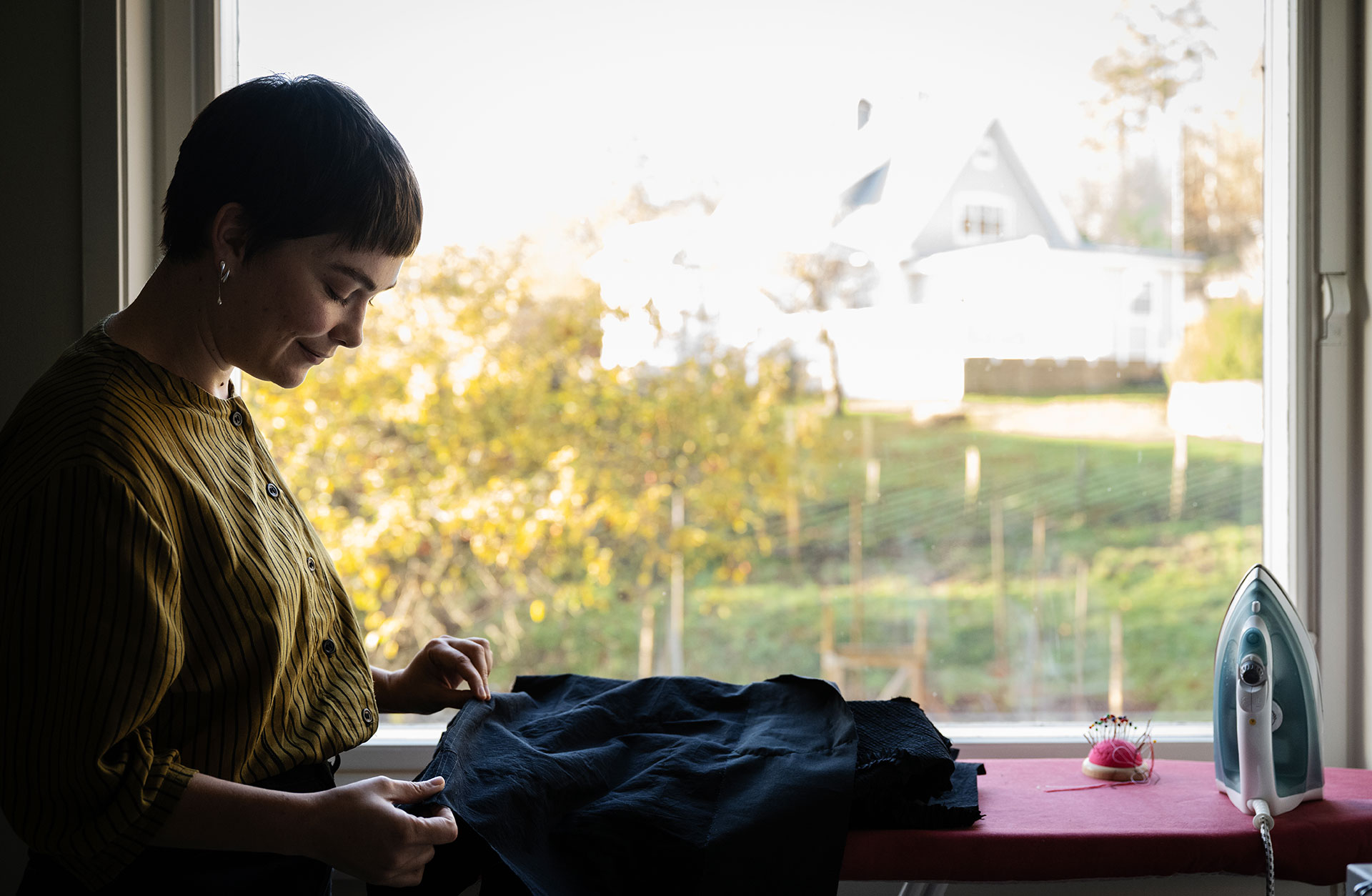 En kvinna med kort frisyr står vid ett fönster och stryker ett plagg.