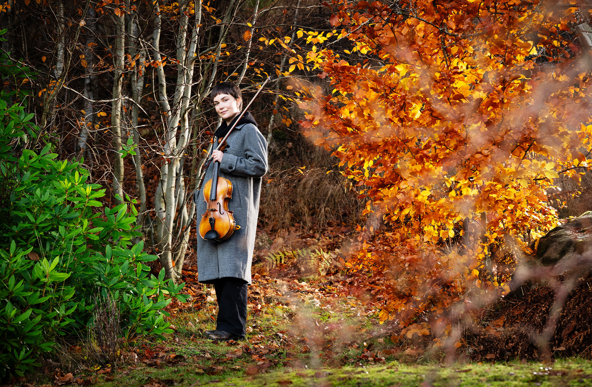 En korthårig kvinna står utomhus. Det är höst med vackra gula löv. Hon är klädd i kappa med ett fiolfodral över axeln.