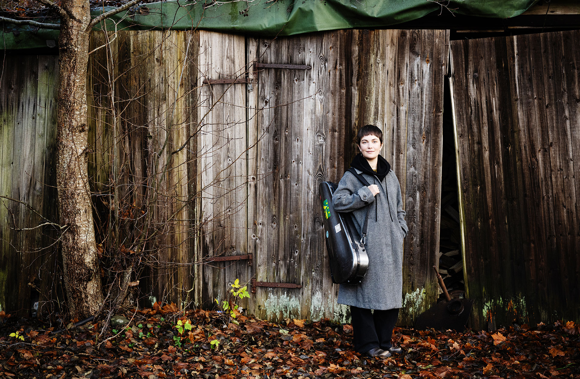 En korthårig kvinna står utomhus framför ett skjul med en gammal trävägg. Hon är klädd i kappa med ett fiolfodral över axeln. Det är höst.