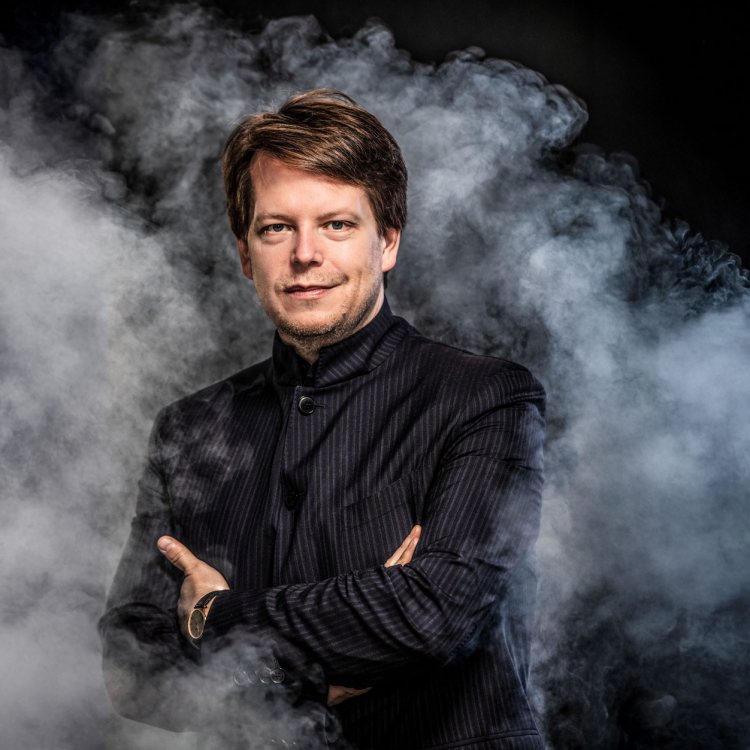 En man i randig mörk kavaj står i ett mörkt rum omgiven av rök.