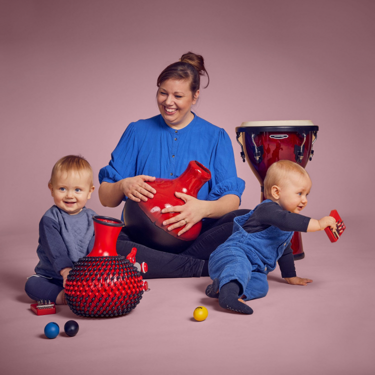 En kvinna sitter med två bebisar runt sig och de leker med instrument. De har blåa kläder mot ljusrosa bakgrund.