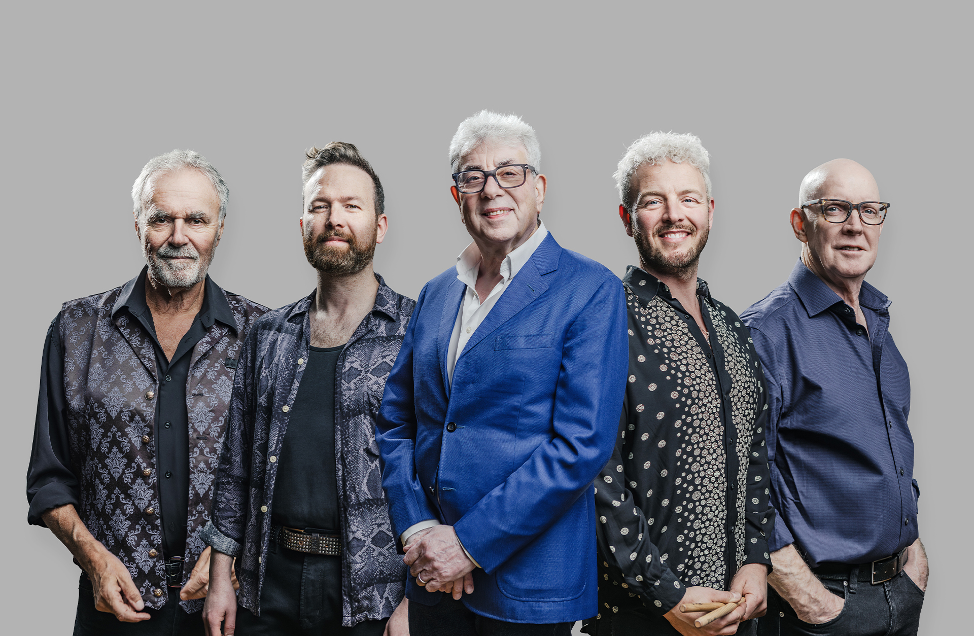 Fem vuxna personer står bredvid varandra framför en neutral grå bakgrund. De är klädda i stilrena, semi‑formella kläder i olika färger och mönster. Personen i mitten bär en klarblå kavaj, medan de andra har skjortor i mönstrat eller enfärgat tyg. Gruppen poserar rakt mot kameran i ett professionellt gruppfoto.