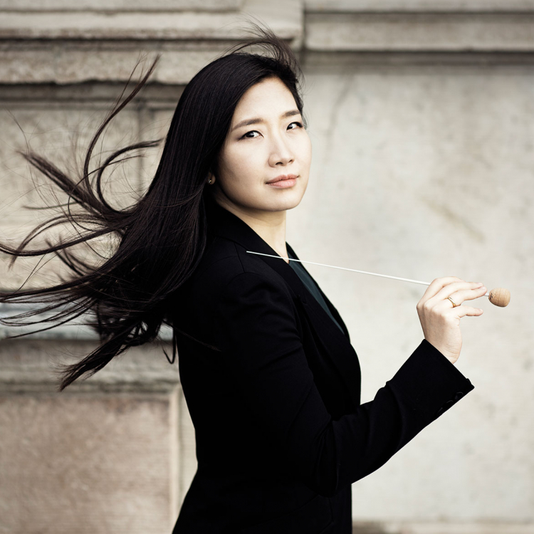 Kvinna med mörkt hår som flyger i vinden, med en taktpinne i handen.