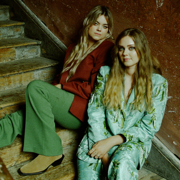 Två kvinnor sitter tillbakalutade i en trappa. De är blonda, med långt hår och de tittar in i kameran.