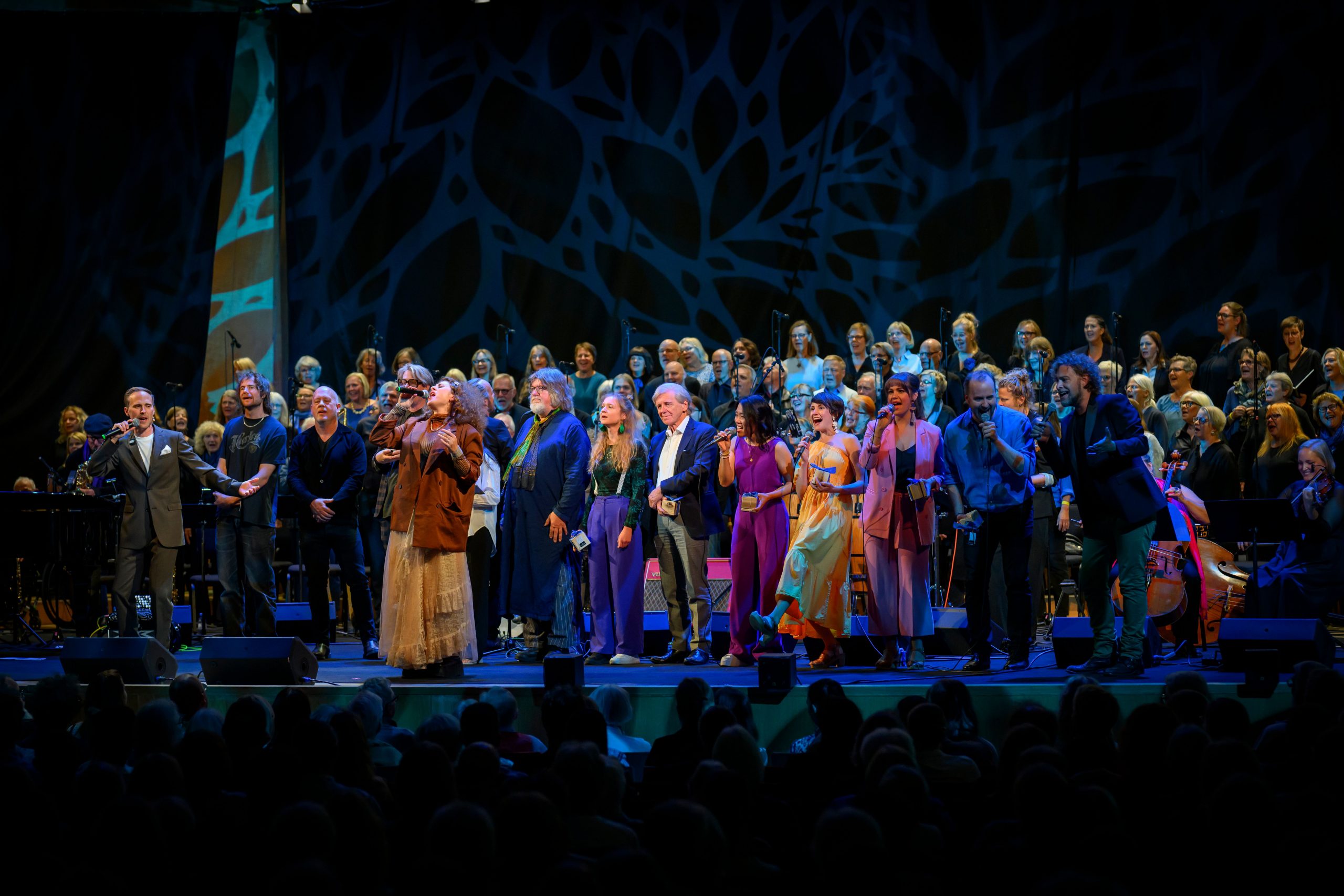 En stor grupp artister står på en upplyst scen under en livekonsert. I förgrunden sjunger flera solister i färgstarka och eleganta kläder, medan en stor kör står bakom dem. Scenbakgrunden består av ett blått, organiskt mönster och publiken syns som siluetter längst fram.