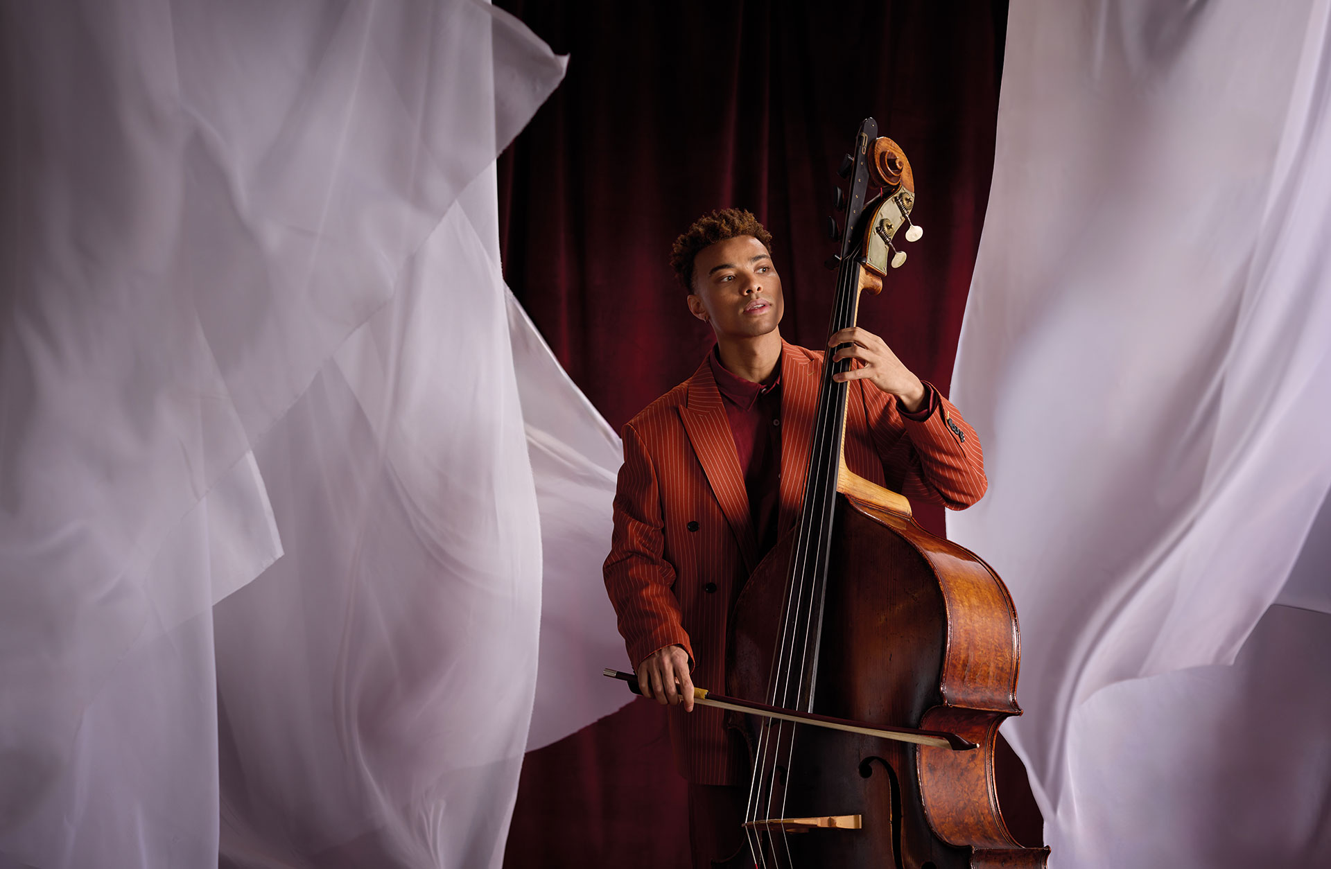 Man med kontrabas i miljö med vita tyger mot röd bakgrund.