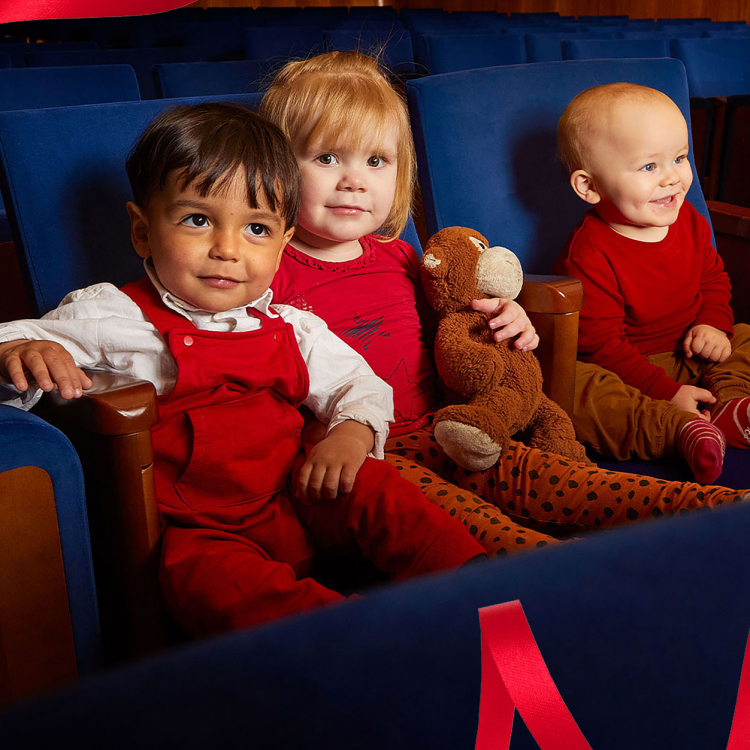 Tre barn sitter i blåa konsertstolar och tittar in i kameran och ler. Det ena barnet håller en nalle i armarna.