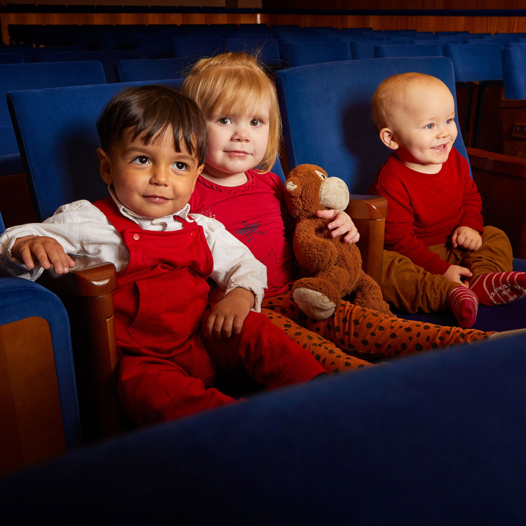 Tre glada små barn i åldern 1-2 år sitter som publik i Stenhammarsalens blå stolar.