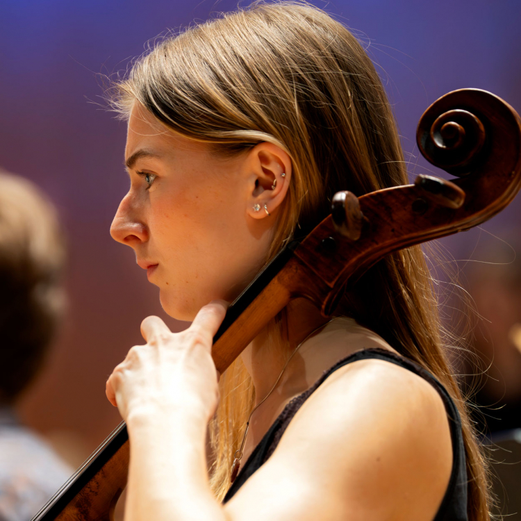Närbild av en ung kvinna med långt hår som spelar cello med en orkester.
