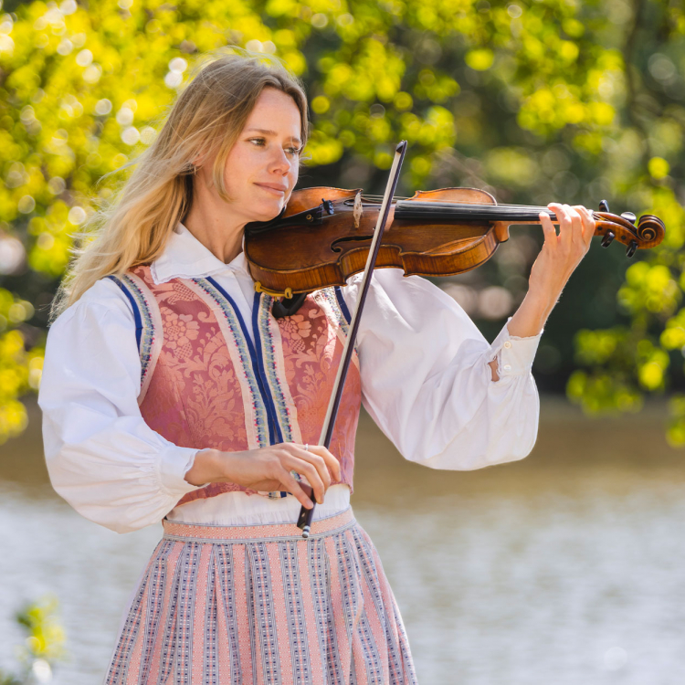 Kvinna står i folkdräkt vid vatten och spelar violin. Grönskande träd i bakgrunden.