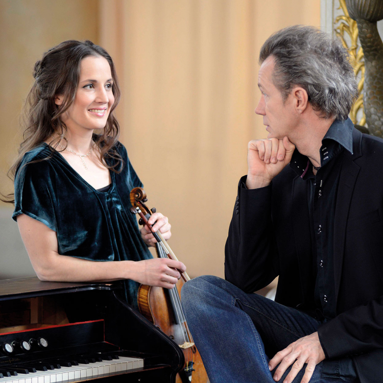 En kvinna håller en fiol i handen, lutar sig mot en orgel och tittar på en man med ryggen mot betraktaren.