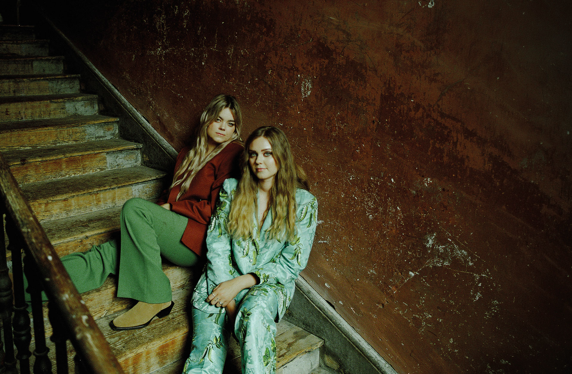 Två kvinnor sitter tillbakalutade i en trappa. De är blonda, med långt hår och de tittar in i kameran.
