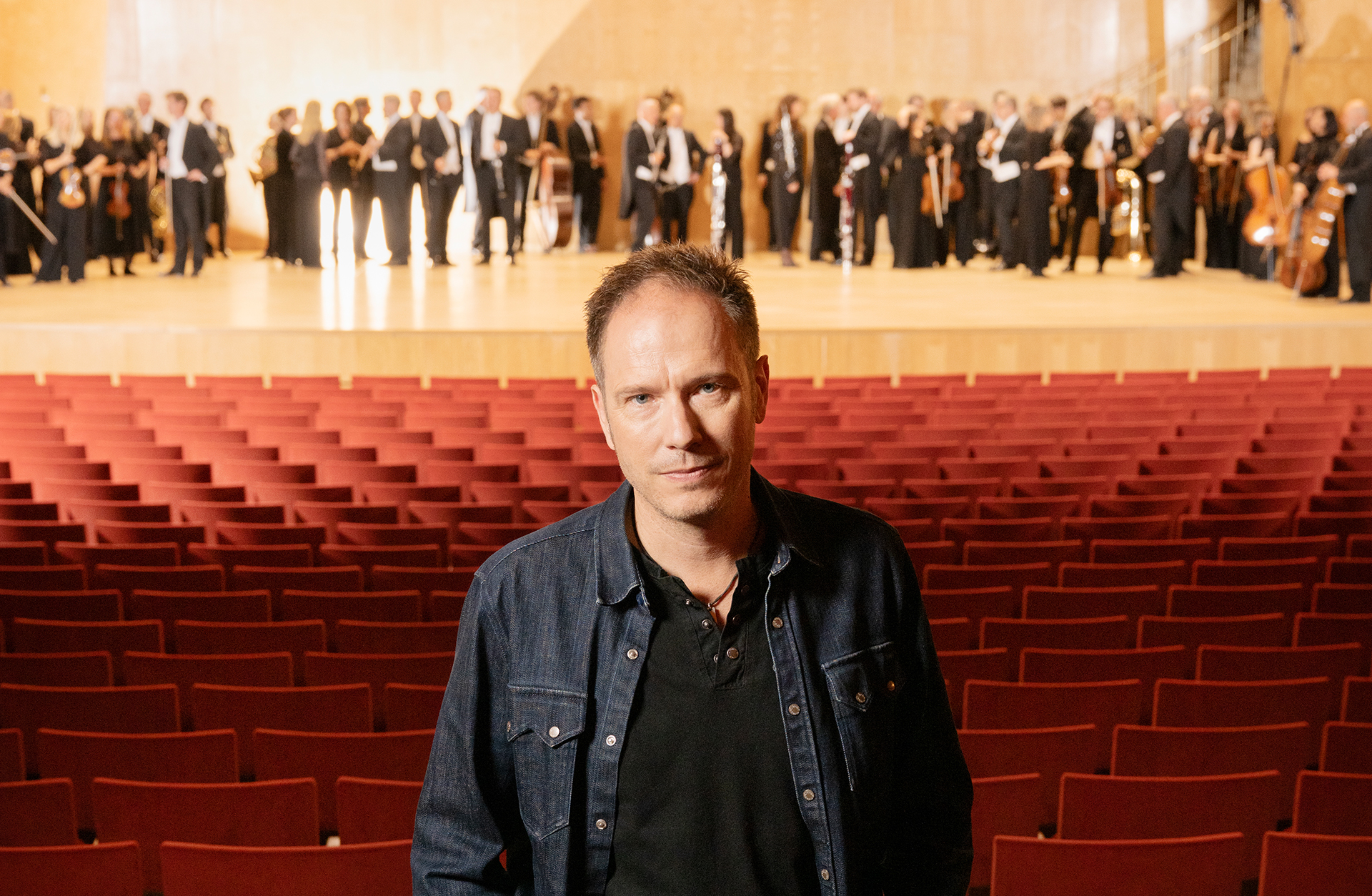 En man står i konsertsalen bland de tomma stolarna på parketten. I bakgrunden ser man en symfoniorkester på scenen.