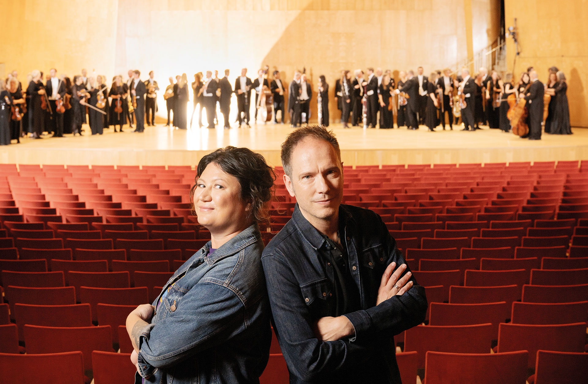 Ina Lundström står rygg mot rygg med Martin Schaub i jeanskläder bland röda salongsstolar med orkestermusiker i bakgrunden.