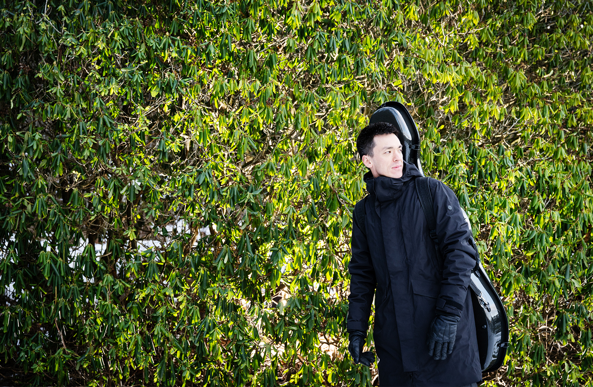 En man i vinterrock står vid en grönbladig buske. Han bär sitt cellofodral på ryggen.