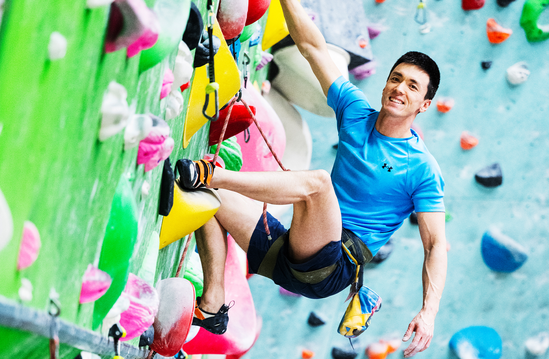 En glad man på en färgglad klättervägg hänger ut från väggen och ler mot kameran.