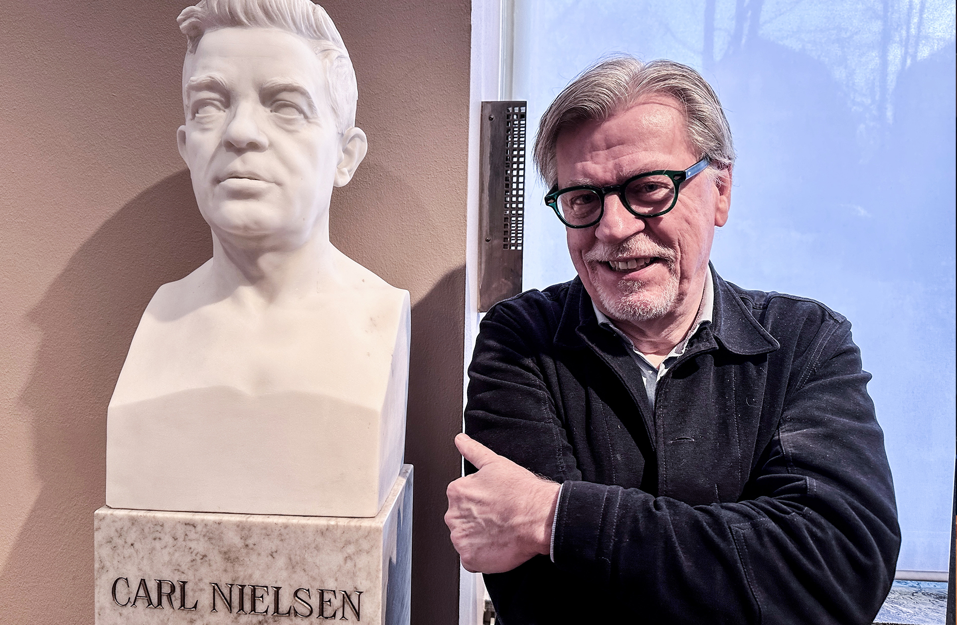 En äldre gråhårig man tittar piggt in i kameran och ler.