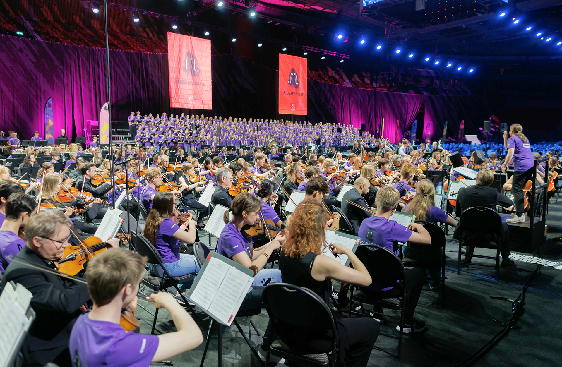 En stor orkester med unga musiker och en kör i bakgrunden och en dirigent i konsert i Scandinavium. Alla har lila tröjor.
