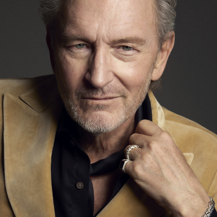 En man i svart skjorta och beige kavaj framför en svart bakgrund 