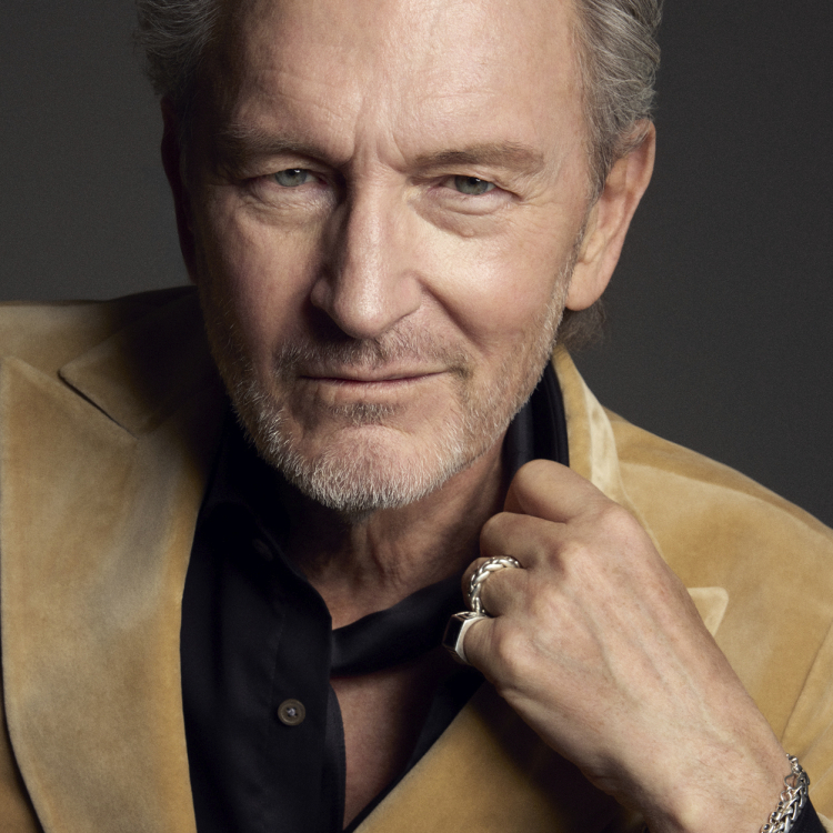 En man i svart skjorta och beige kavaj framför en svart bakgrund