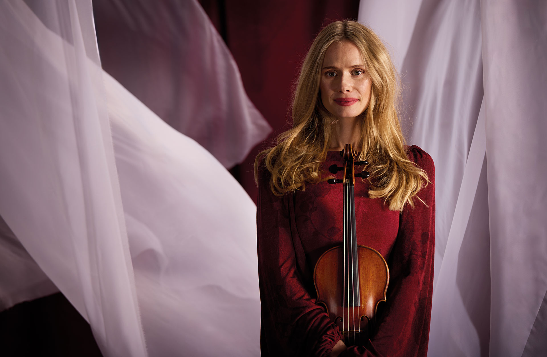 Violinisten Tove Lund med fiolen i famnen, fotad mot röd bakgrund med vita böljande tyger.