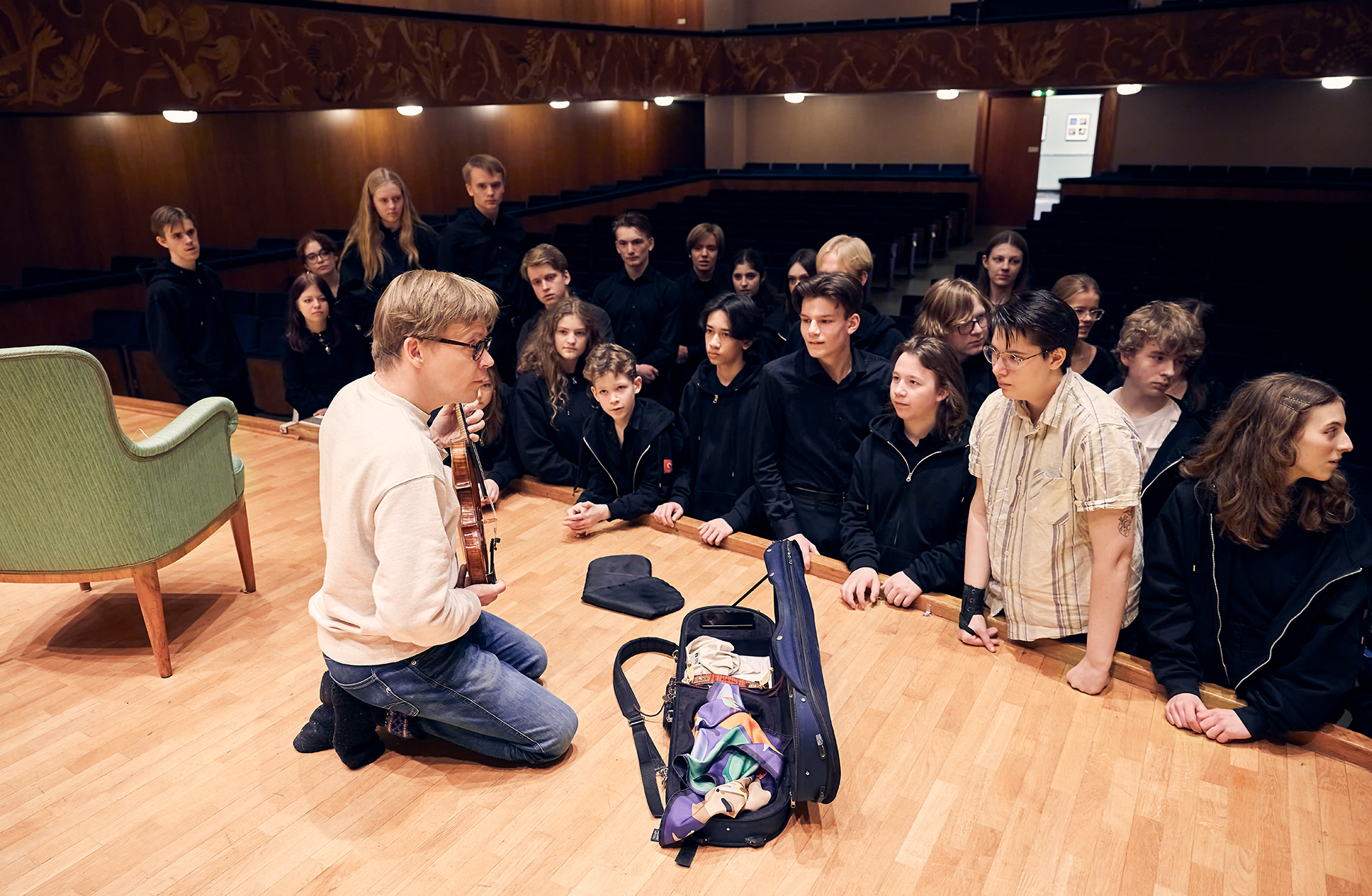 En blond kortklippt man med jeans och collegetröja sitter på knä vid scenkanten med sin fiol i handen. Han visar den för en skara intresserade ungdomar som lutar sig fram över scenkanten.