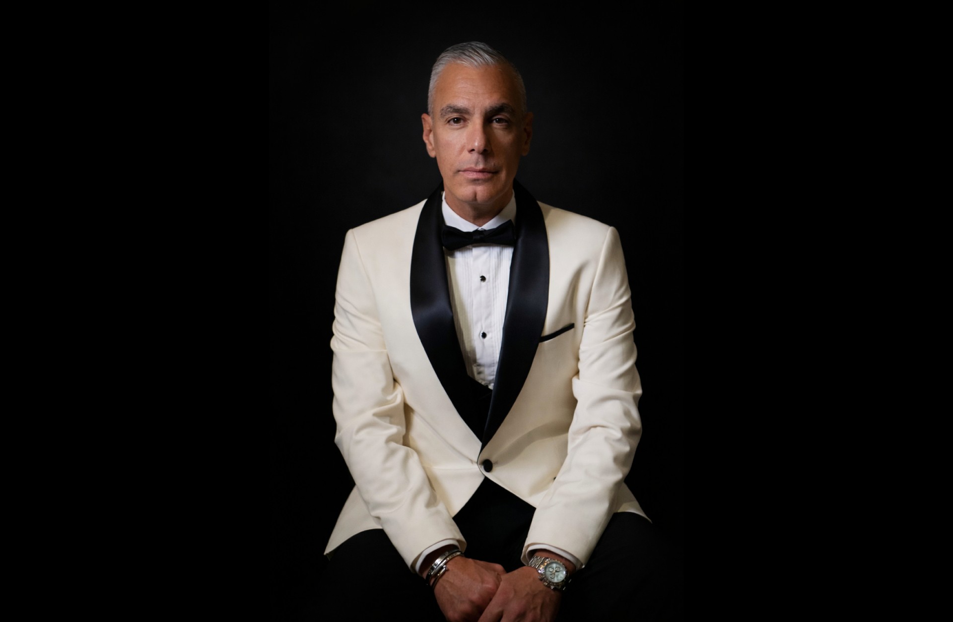 En man sitter framför en svart vägg, han har en vit skjorta, svart fluga och en vit kavaj med svarta slag.