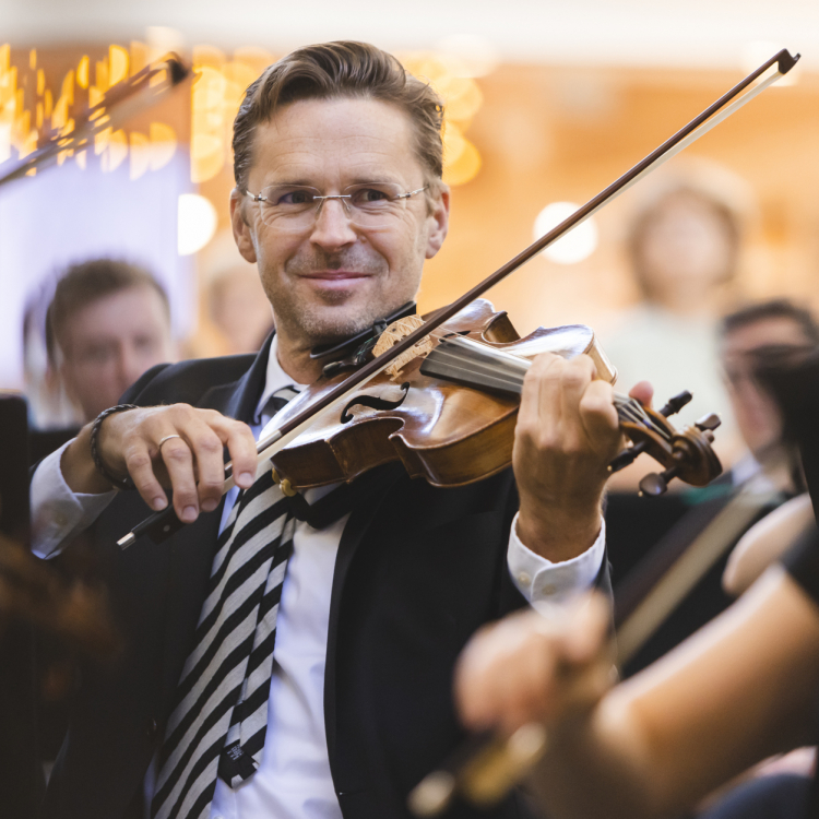 En glad man, klädd i svart kavaj och randig slips, tittar in i kameran och ler. Han spelar violin.