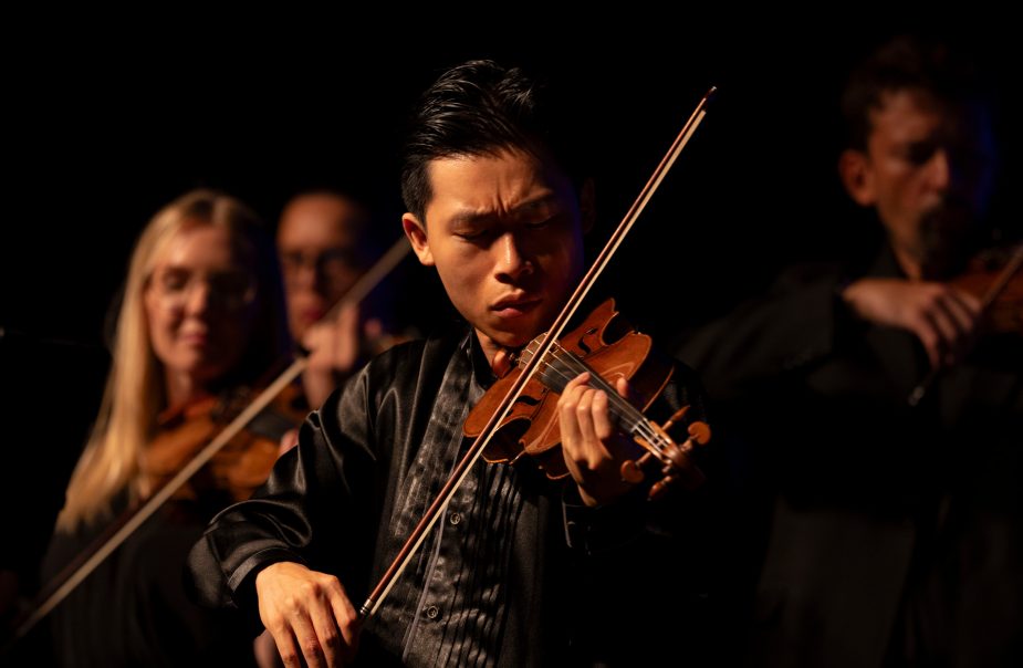 En violinist upplyst på scen medan andra musiker spelar i bakgrunden.