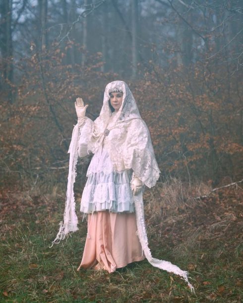 En person i ett dimmigt skogslandskap, klädd i lager av ljusa, vintageinspirerade tyger, står med ena handen lyft som i en hälsning.