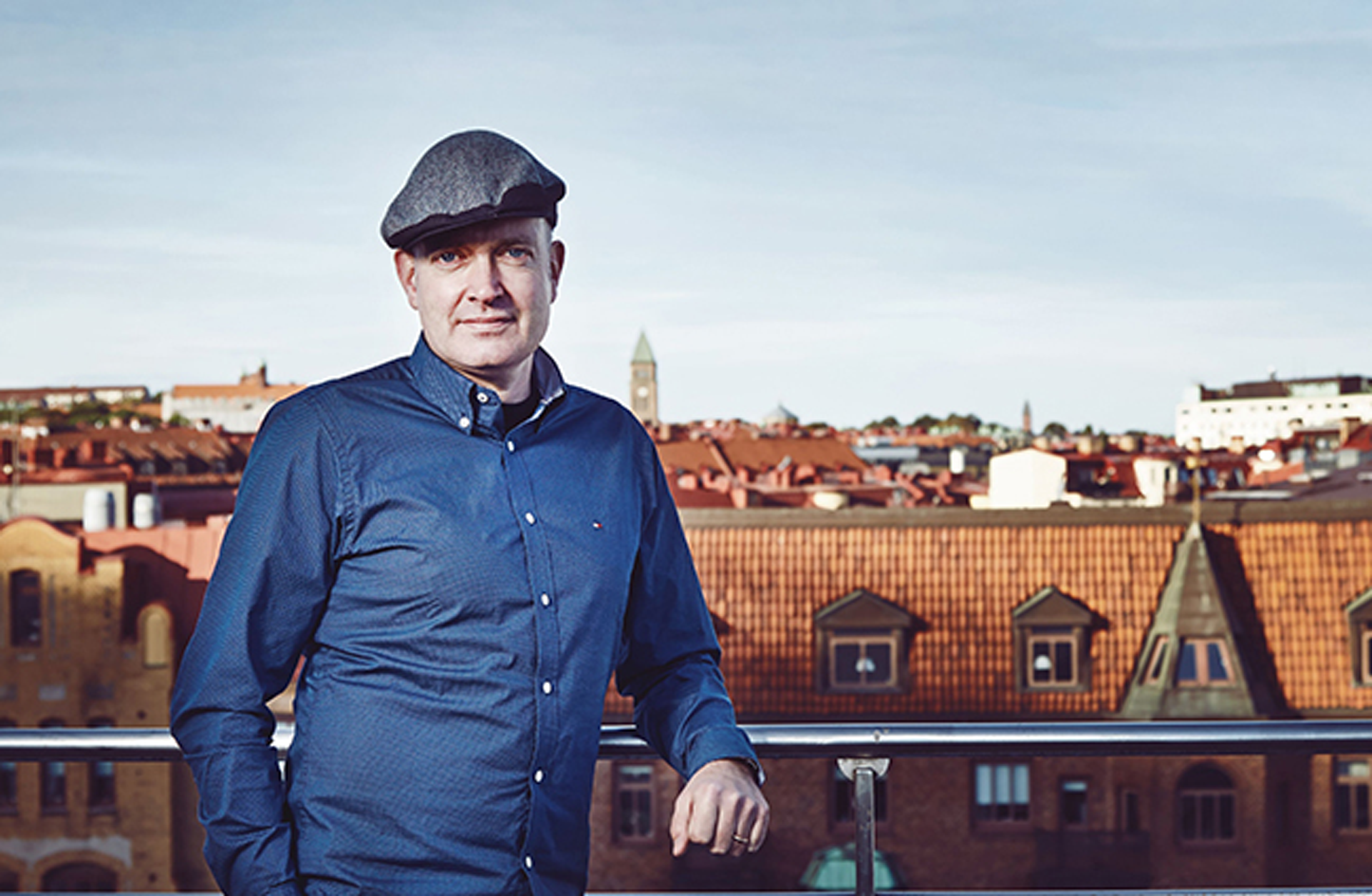 En man i blå skjorta och keps står på en balkong. I bakgrunden ser man takåsar mot en blå himmel.