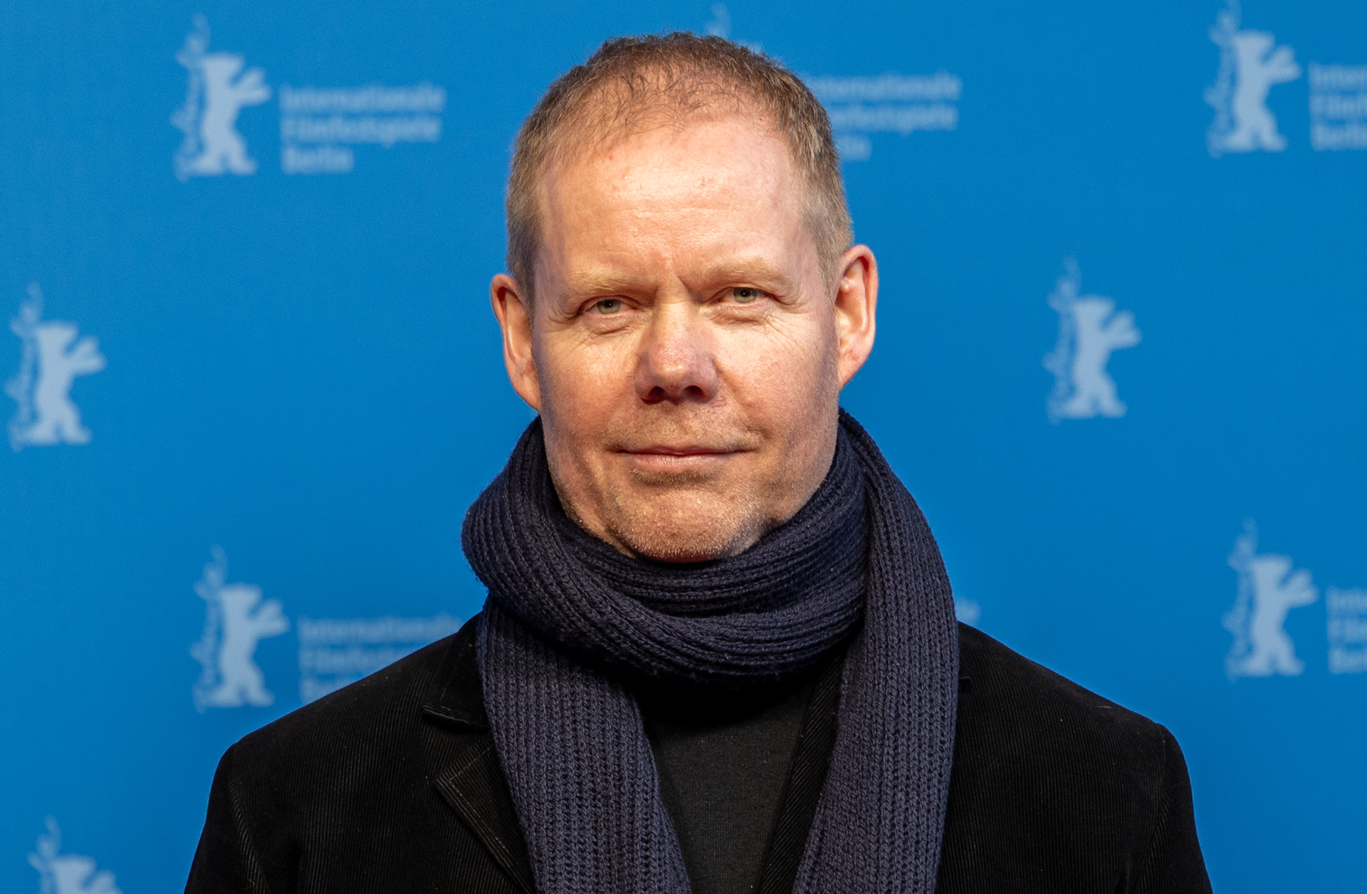 Max Richter, står mot en blå vägg. Han tittar in i kameran och ler med stängd mun.