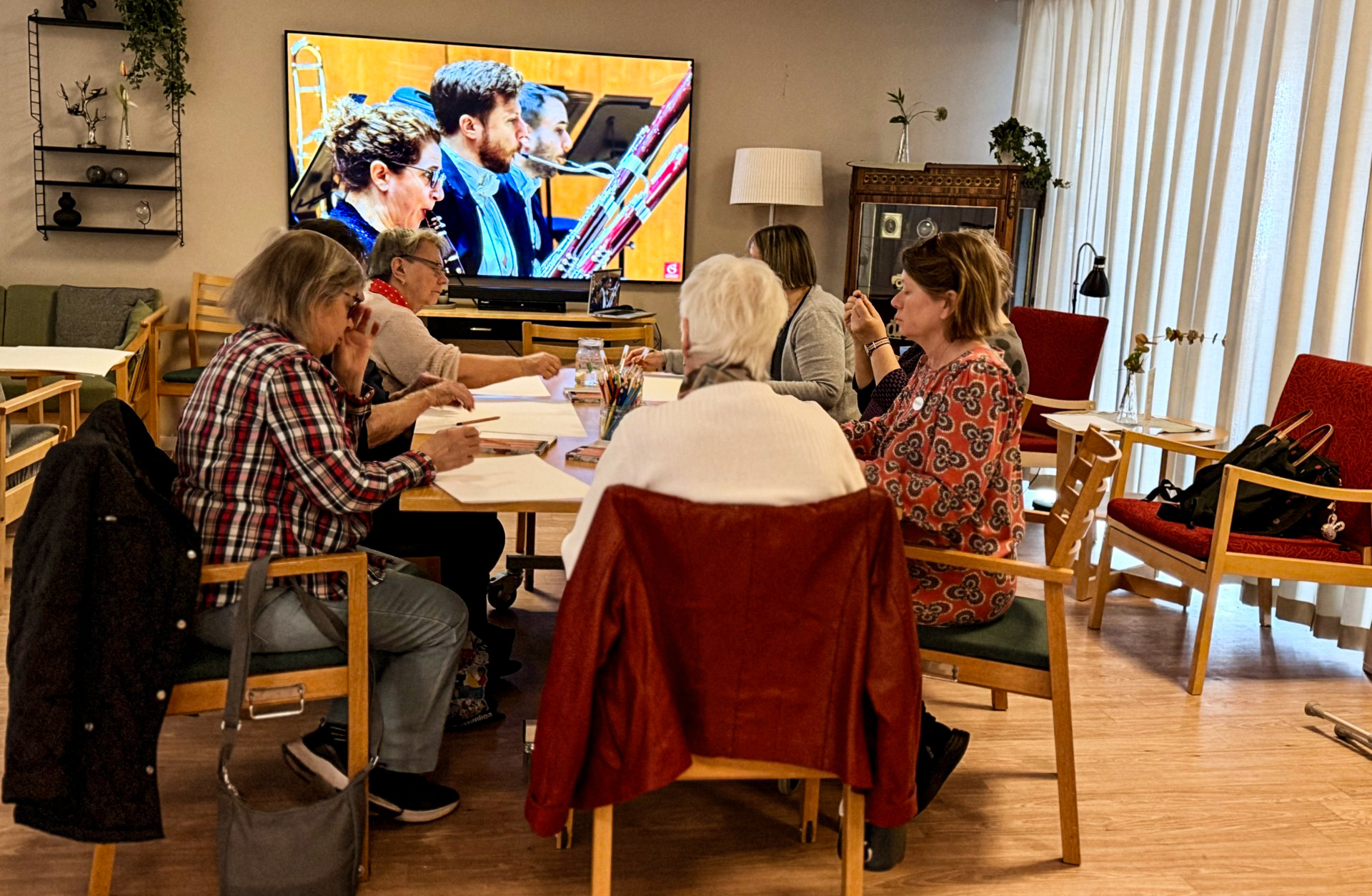 En grupp äldre människor sitter runt ett bord. På en tv ser man musiker i närbild.