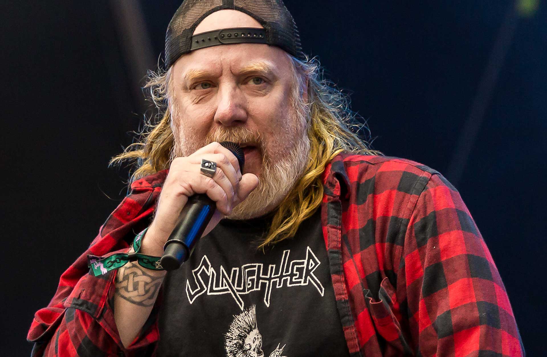 Bild av en äldre man i långt hår och skägg som sjunger i en mikrofon. Han har en bakåtvänd keps på huvudet och är klädd i en svart t-shirt med ett rock-tryck och en rödrutig skjorta.